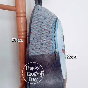 May include: A blue and gray patchwork backpack with a zipper closure and a Happy Quilt Day logo. The backpack measures 28 cm tall, 22 cm wide, and 11 cm deep.
