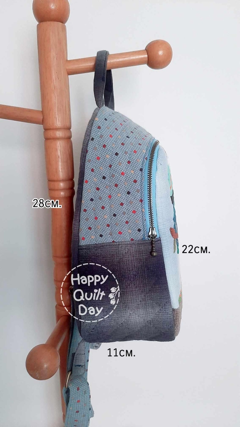 May include: A blue and gray patchwork backpack with a zipper closure and a Happy Quilt Day logo. The backpack measures 28 cm tall, 22 cm wide, and 11 cm deep.