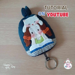 May include: A blue fabric keychain featuring a girl and a cat design. The girl has brown hair and is wearing a green shirt. The cat is white and is sitting on the girl's head. The keychain has a silver ring and a loop for attaching to keys. The text "TUTORIAL ON YOUTUBE" is written in white letters on a blue background. The text "Happy quilt Day" is written in white letters on a pink background. The text "2021 NOV.20" is written in white letters on a pink background.