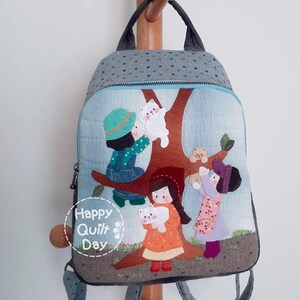 May include: A gray and blue backpack with a quilted design featuring a scene of children playing with cats in a tree. The backpack has a zipper closure and adjustable straps. The text "Happy Quilt Day" is printed on the front of the backpack.