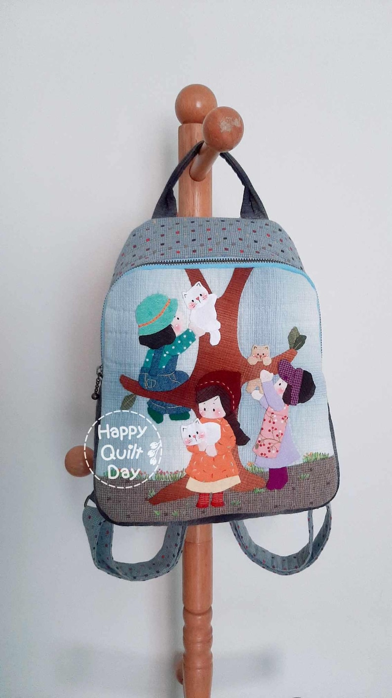 May include: A gray and blue backpack with a quilted design featuring a scene of children playing with cats in a tree. The backpack has a zipper closure and adjustable straps. The text "Happy Quilt Day" is printed on the front of the backpack.