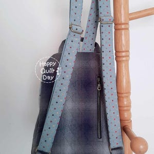 May include: A blue and gray backpack with a zippered pocket and adjustable straps with a white and red polka dot pattern. The straps are attached to a wooden rack.