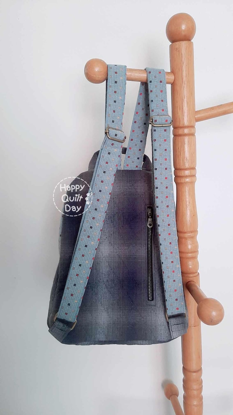 May include: A blue and gray backpack with a zippered pocket and adjustable straps with a white and red polka dot pattern. The straps are attached to a wooden rack.