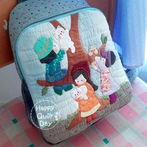 May include: A blue quilted backpack with a tree and children applique. The backpack has a zipper closure and a top handle. The text "Happy Quilt Day" is on the front of the backpack.