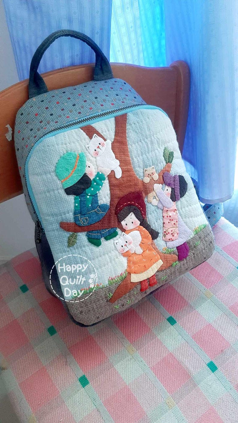 May include: A blue quilted backpack with a tree and children applique. The backpack has a zipper closure and a top handle. The text "Happy Quilt Day" is on the front of the backpack.