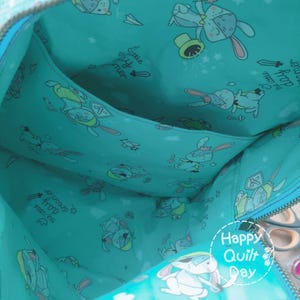 May include: A turquoise fabric bag with a zipper closure and a pocket on the inside. The fabric has a repeating pattern of bunnies wearing knight costumes and the text "Little knighty timer" and "The happy troop is awesome". The bag has a Happy Quilt Day logo on the bottom.