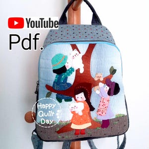 May include: A blue and grey quilted backpack featuring a design of children playing with cats in a tree. The backpack has a zip fastening and adjustable straps. The text "Happy Quilt Day" is on the front of the backpack.