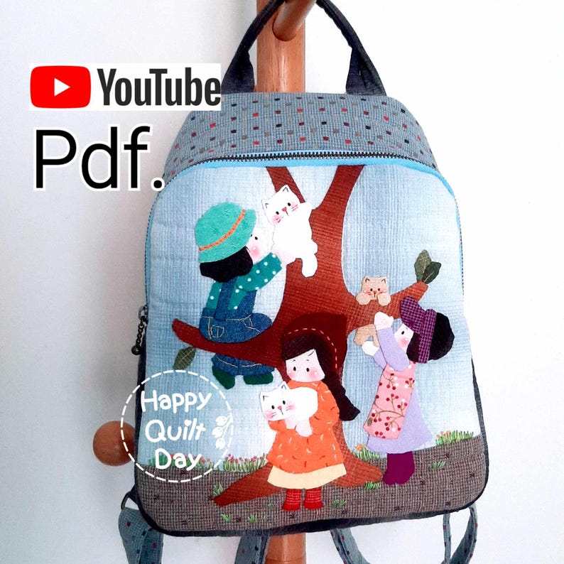 May include: A blue and gray quilted backpack with a design of children playing with cats in a tree. The backpack has a zipper closure and adjustable straps. The text "Happy Quilt Day" is on the front of the backpack.