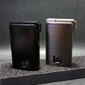 May include: Two rectangular lighters, one black and one gunmetal, are displayed side-by-side. The lighters have a sleek, minimalist design with a textured finish. The base of each lighter features a rectangular cutout.