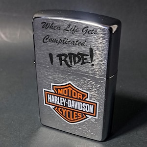 May include: A silver-toned Harley-Davidson lighter with the text "When Life Gets Complicated... I RIDE!" The Harley-Davidson logo is displayed in orange and white. The lighter has a brushed metal finish.