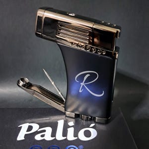 May include: A black and silver Palio Pro lighter with a curved design and the letter "R" in a script font. The lighter is on top of a box with the Palio Pro logo. The lighter has a built-in cigar punch and a flame adjustment.