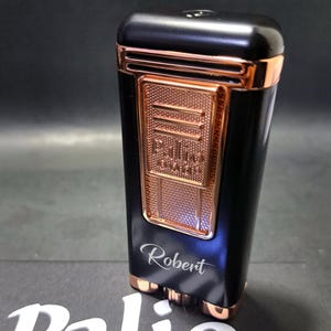 Op de afbeelding: Zwart en koperkleurig Palio aansteker met de naam "Robert" gegraveerd. De rechthoekige aansteker heeft een koperkleurig paneel met textuur en accenten. Het woord "Palio" is zichtbaar.