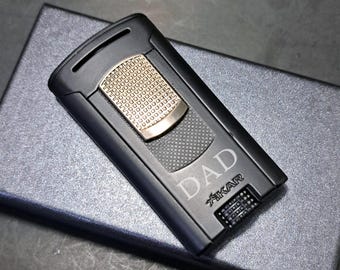 May include: A black cigar lighter with a gold-coloured accent and the word "DAD" engraved on the front. The lighter is rectangular and rests on a dark grey surface. The brand name "XIKAR" is also visible.