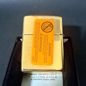 May include: Gold-colored Zippo lighter with a warning label in English and Spanish. The label states to keep the lighter away from children and to read instructions before using. The text below the lighter reads "Armor. The Armor case is about 1.5 times as thick as a standard Zippo case. The Armor logo."