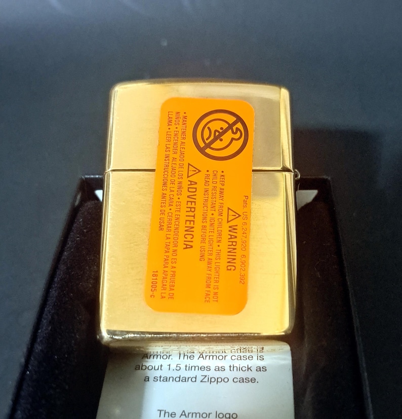 May include: Gold-colored Zippo lighter with a warning label in English and Spanish. The label states to keep the lighter away from children and to read instructions before using. The text below the lighter reads "Armor. The Armor case is about 1.5 times as thick as a standard Zippo case. The Armor logo."