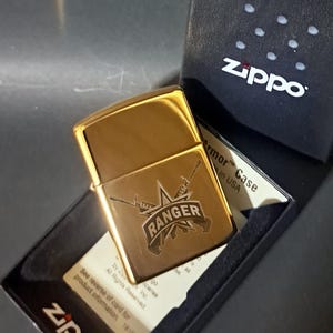 May include: Gold Zippo lighter with an engraved design of a star, two rifles, and the word "RANGER". The lighter is in a black box with the Zippo logo.
