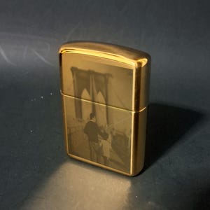 May include: A gold-colored lighter with an engraved image of the Brooklyn Bridge and a couple walking across it.