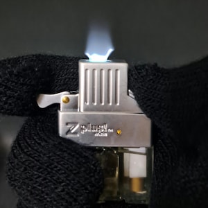 May include: A silver Z-plus butane lighter insert with a vertical ribbed top section and a clear plastic fuel tank base. The lighter is actively emitting a bright blue flame from the top nozzle, with the brand name Z-plus printed on the side.
