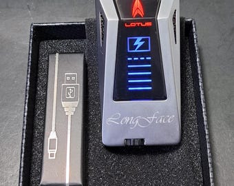 May include: A silver Lotus B2 lighter with a red logo and blue display, presented in a black box. The lighter features the text "Long Face" and includes a USB charger. The box also lists features like triple torch flames and a fold-out cigar punch.