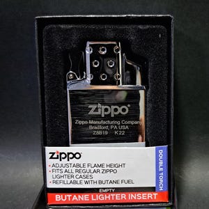 May include: A Zippo butane lighter insert in a black box. The silver-colored insert features the Zippo logo and text. The packaging includes text that reads "Adjustable Flame Height," "Fits All Regular Zippo Lighter Cases," and "Refillable with Butane Fuel."