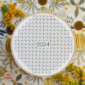 May include: A white embroidery hoop with a black grid pattern and the number 2024 in the center. The hoop is on a floral fabric background.