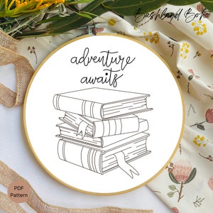 May include: A black and white line drawing of a stack of books with the text "adventure awaits" above the books. The image is on a white background and is in a wooden embroidery hoop.