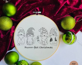 May include: A white embroidery hoop with a black and white line drawing of four cats wearing Christmas hats. The text "Purrrr-fect Christmas" is written below the cats. The hoop is surrounded by green Christmas ornaments and a red velvet background.