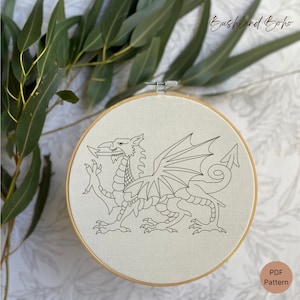 May include: A black and white line drawing of a dragon on a white fabric stretched over a wooden embroidery hoop. The text "Bushland Boho" is in the upper right corner of the image. The text "PDF Pattern" is in the lower right corner of the image.