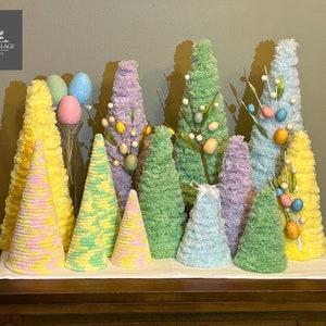 May include: A collection of colorful, cone-shaped decorative trees in shades of yellow, green, blue, and purple. Some trees are adorned with small, decorative eggs and sprigs. The arrangement includes a vase with pastel-colored Easter eggs. The logo "Poppy's Village Home Decor" is in the upper left.