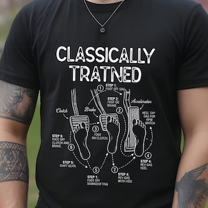 Może przedstawiać: Czarny t-shirt z krótkim rękawem z białym nadrukiem graficznym przedstawiającym schemat pedałów samochodowych oraz napis "CLASSICALLY TRATNED". Projekt ilustruje ułożenie stóp na sprzęgle, hamulcu i gazie z ponumerowanymi krokami.