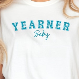 Op de afbeelding: Een wit T-shirt met korte mouwen en het woord YEARNER in een verweerd, blauwgroen collegiaal lettertype in een boog over de borst, met het woord Baby eronder in een kleiner, cursief blauwgroen lettertype. Het shirt heeft een ronde hals.