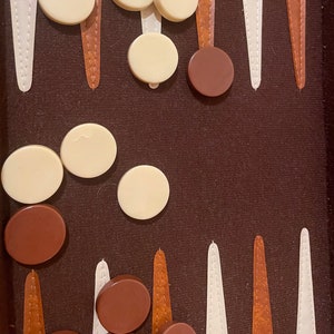 May include: A brown backgammon board with white and brown pieces. The board has a brown felt surface with white and brown lines. There are 10 white pieces and 3 brown pieces on the board.