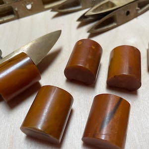 May include: A collection of smooth, cylindrical amber-coloured buttons with marbled brown patterns. Several buttons are arranged in a group, while one button is attached to a metallic brass-coloured tool with a pointed end and a small screw.