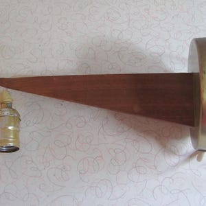 May include: A mid-century modern wall sconce with a wooden arm and a brass-colored metal base. The sconce features a light socket and a white electrical cord. The wooden arm is triangular in shape and extends from the base.