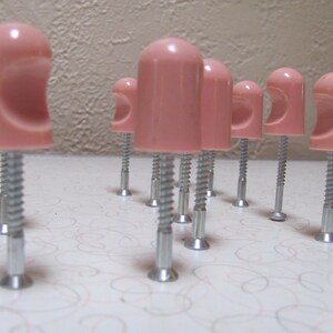 May include: A collection of pink plastic screw covers with a curved design, attached to silver screws. The objects are arranged on a patterned surface, with a neutral-toned wall in the background.