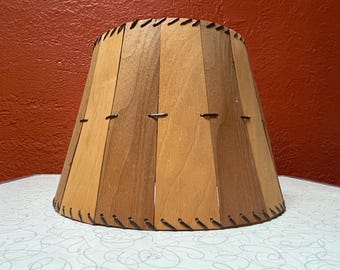 May include: A conical lampshade constructed from alternating light and dark brown wooden panels. The panels are joined together by thin, dark metal wire stitching visible along the top and bottom edges and across the centre of the shade.