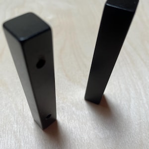 May include: Two matte black cabinet pulls with a square profile and elongated rectangular shape. One pull is positioned vertically showing two circular screw holes on its front face, while the other stands upright beside it, displaying a smooth surface.