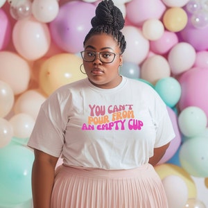 May include: A woman wearing a white t-shirt with the text "YOU CAN'T POUR FROM AN EMPTY CUP" in pink and orange letters. She is standing in front of a wall of pastel balloons.