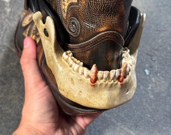 May include: A brown leather cowboy boot featuring a decorative, off-white human jawbone attachment with visible teeth and reddish-brown stained fangs. The boot has intricate embossed swirl patterns and a black leather strap with a metal buckle.