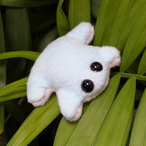 May include: A light blue plush toy shaped like a frog with black eyes. The frog is lying on a bed of green leaves.