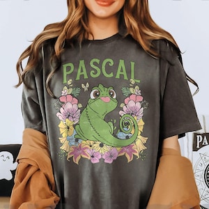 May include: Dark gray t-shirt featuring the name "PASCAL" in green above a cartoon chameleon surrounded by colorful flowers. The chameleon is green with a pink tongue and a curled tail. The shirt has a vintage, washed look.