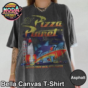 May include: Asphalt-colored Bella Canvas T-shirt featuring a Pizza Planet graphic. The design includes a rocket, a space-themed restaurant, and the text "Pizza Planet" in a retro font. The shirt also has the text "Serving Your Local Star Cluster."