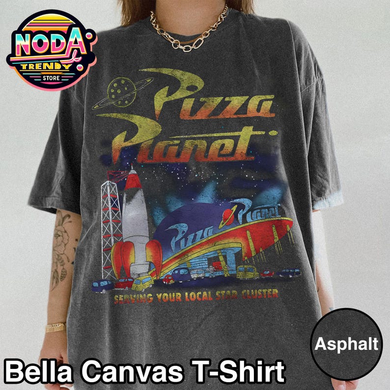 May include: Asphalt-colored Bella Canvas T-shirt featuring a Pizza Planet graphic. The design includes a rocket, a space-themed restaurant, and the text "Pizza Planet" in a retro font. The shirt also has the text "Serving Your Local Star Cluster."