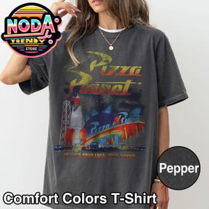 May include: Dark grey Comfort Colors T-shirt featuring a Pizza Planet graphic. The design includes a rocket, a space-themed restaurant, and the text "Serving Your Local Star Cluster". The shirt is a Pepper color.
