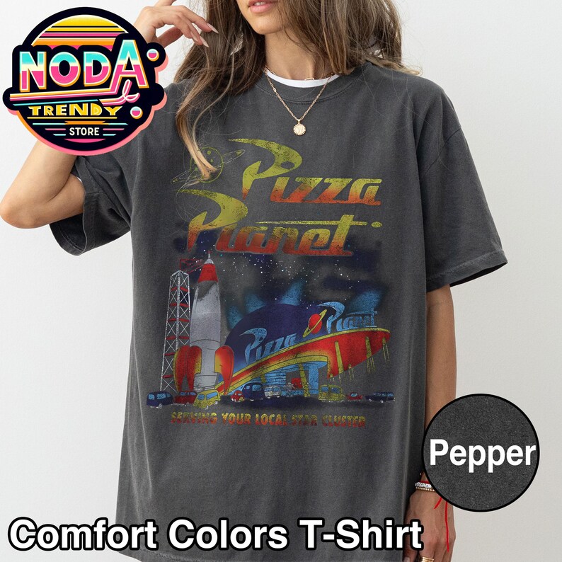 May include: Dark grey Comfort Colors T-shirt featuring a Pizza Planet graphic. The design includes a rocket, a space-themed restaurant, and the text "Serving Your Local Star Cluster". The shirt is a Pepper color.