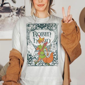 May include: White t-shirt featuring a graphic of Robin Hood, with the text "Robin Hood" in a decorative frame. The design includes illustrations of characters from the animated film. The shirt has short sleeves.