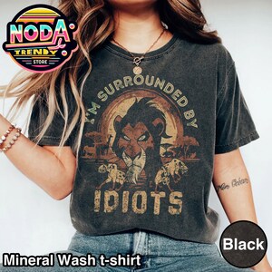 Może przedstawiać: Czarny t-shirt z mineralnym praniem z grafiką lwa i napisem "I'M SURROUNDED BY IDIOTS". Projekt zawiera tło zachodu słońca i sylwetki zwierząt. Koszulka ma wygląd vintage.