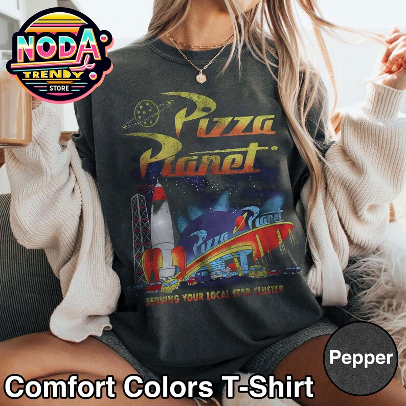 May include: Dark gray Comfort Colors T-shirt featuring a Pizza Planet graphic with a rocket, planet, and the text "Serving Your Local Star Cluster." The shirt also has the text "Pizza Planet" in yellow and red.