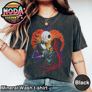 以下が含まれることがあります： 黒色のミネラルウォッシュTシャツ。映画「ナイトメアー・ビフォア・クリスマス」のジャック・スケリントンとサリーのグラフィックが、ハート型のオレンジと紫の背景に描かれています。Tシャツには「Mineral Wash t-shirt」と「Black」の文字が入っています。