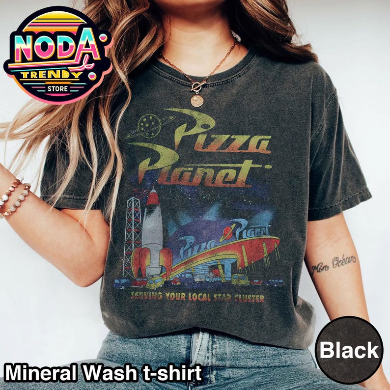 May include: Black mineral wash t-shirt featuring the Pizza Planet logo with a rocket ship and the text "SERVING YOUR LOCAL STAR CLUSTER." The shirt has a vintage, distressed look.